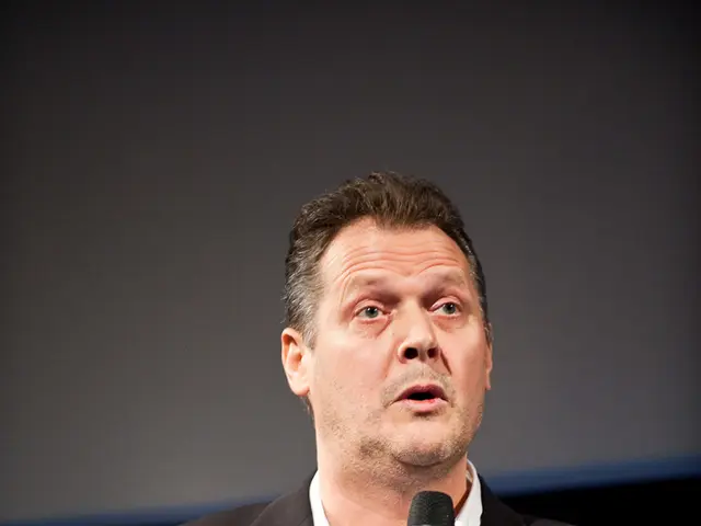 As we can see in the image, there is a man wearing black color jacket and talking on mike.