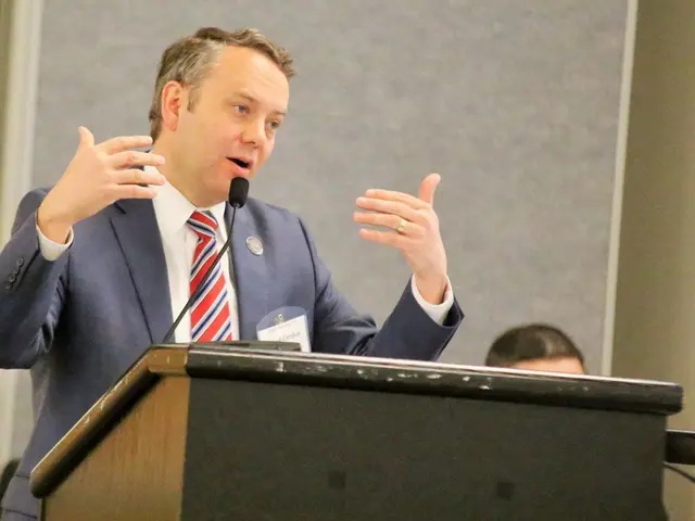 In this image there are people, podium, mic, wall and objects. In-front of that man there is a...