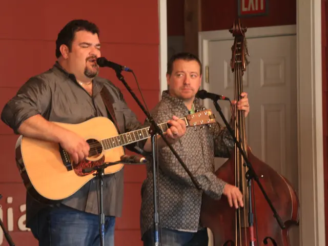 A man singing with mic in front of him playing a guitar. Another man beside him is playing cello.