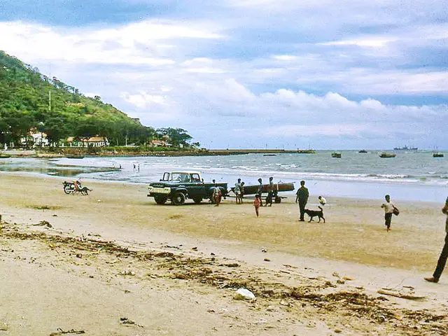 In this image there are people walking on a beach and there is a vehicle, in the background there...