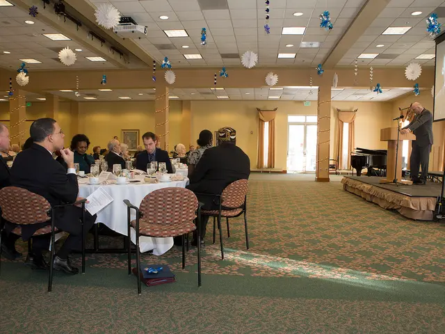 A man is standing at a podium on a stage. There is a screen behind him. People are sitting in...