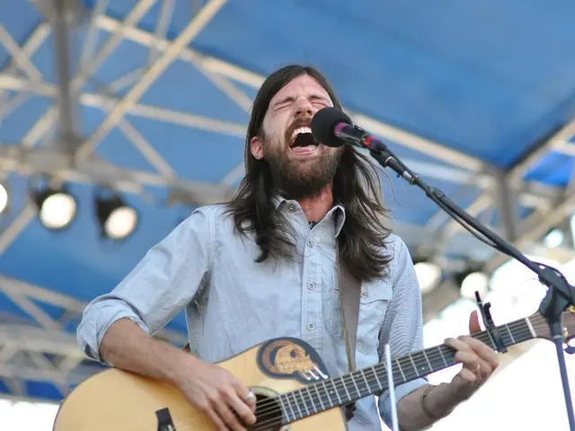 In the image there is man with long hair singing on mic playing guitar and above its stage, poles...