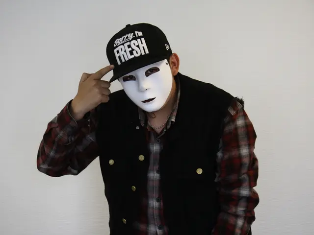 This image consists of a man wearing a mask along with a black jacket and a cap. In the background,...