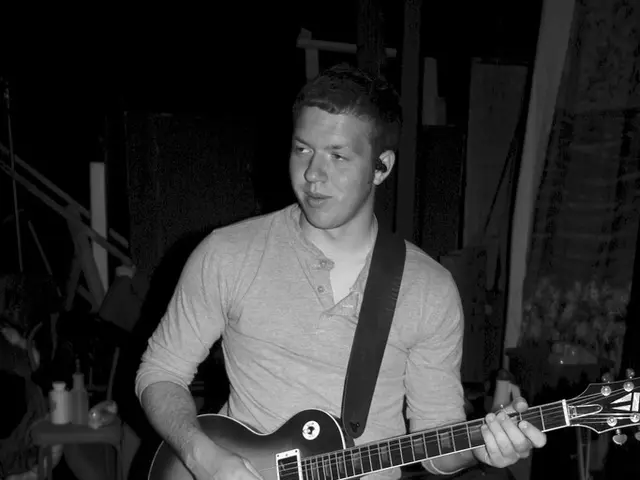 In this image this man is standing and playing the guitar which is in hands. In the background...