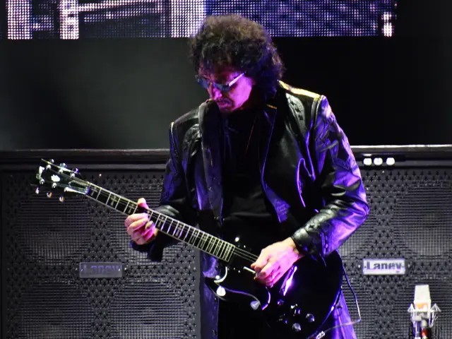 In this image I see a man who is wearing a jacket and is holding the guitar. In the background I...