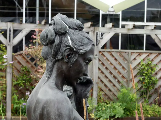 This picture shows statue of a woman. We see plants and a wooden fence.