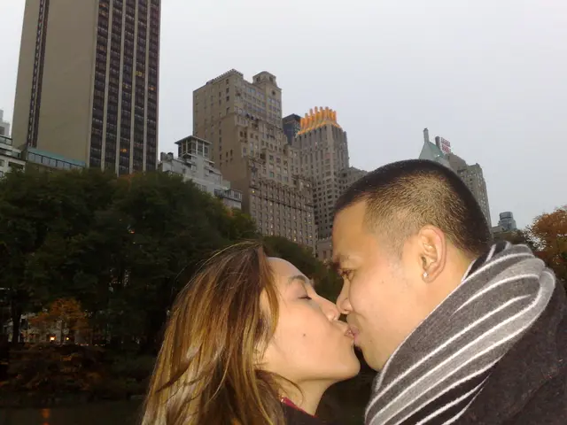 In this image I see a man and a woman who are kissing and in the background I see the trees,...