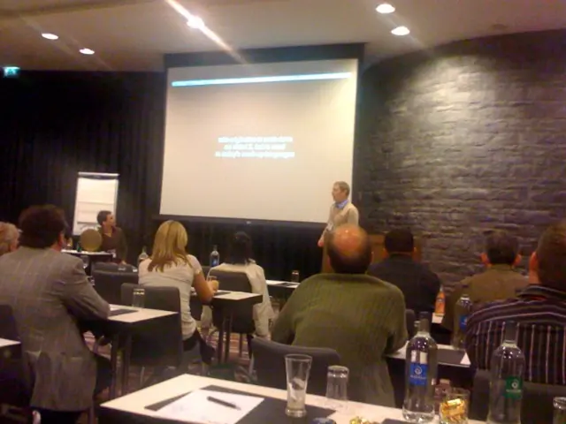 A group of people are listening to a man whose is presenting with a screen behind him.