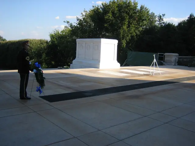 This picture shows a man standing and holding a stand with flowers to it, Man wore a cap on his...