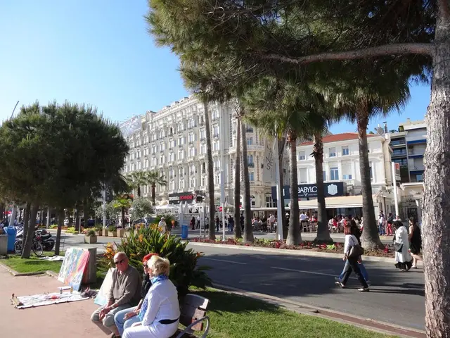 This is a picture of a city , where there are three persons sitting on the bench , plants, grass,...