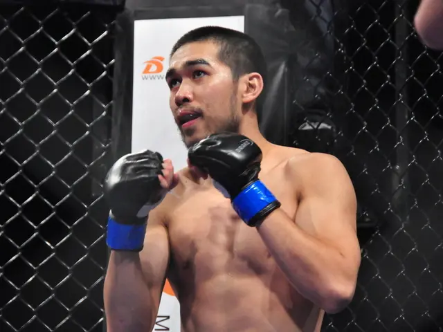 in this image there is a man standing. He is wearing boxing gloves. Behind him there is a fence....