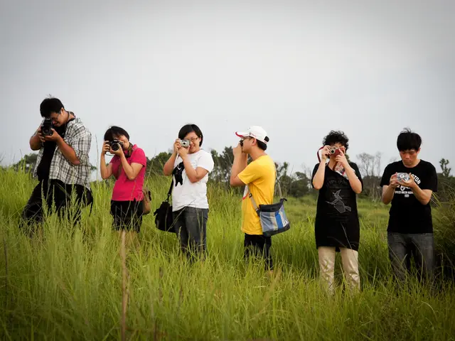 In the foreground I can see six persons are standing on the grass and are holding cameras in their...