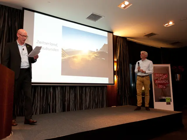 In this image there are 2 men standing on the stage. The person on the right is holding a...