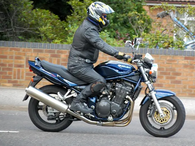 In this image a person wearing a black jacket is sitting on the bike and riding on the road. Behind...