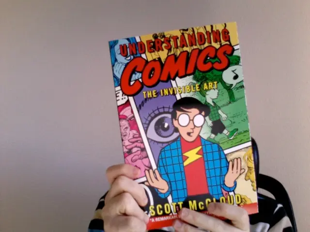 In this picture, we see the hands of a man is holding a comic book which is in blue, pink, green,...