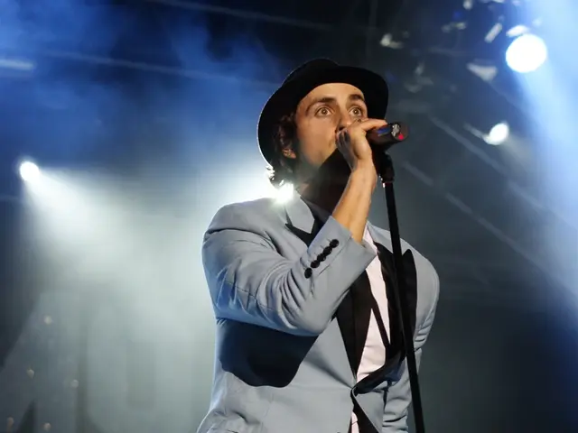 In this image there is a person singing on a mike. He is wearing a grey blazer, grey trousers and a...