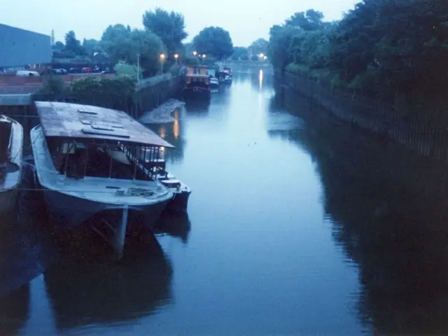 In the picture there are many boats floating beside a land and there are few vehicles parked in the...