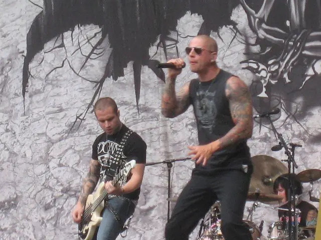 A person wearing a black shirt and black pants is singing and holding a mic. Behind him another...