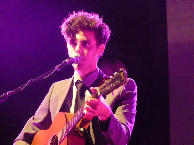 In this picture, we see a man is wearing the grey blazer. He is holding a guitar in his hands and...