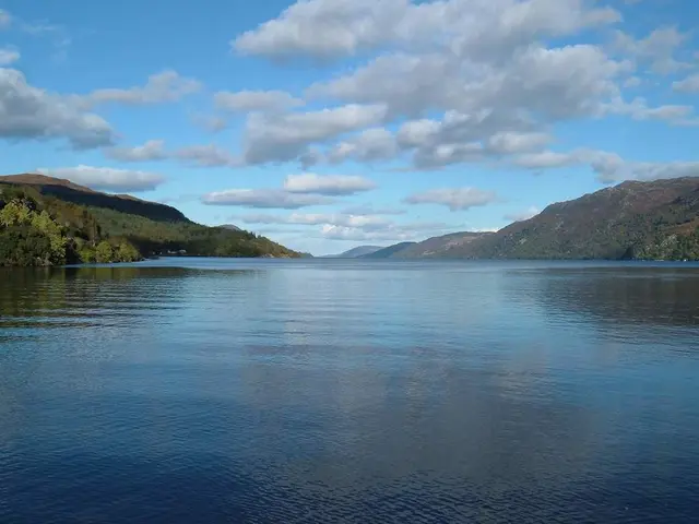 In this image there is water, hills, trees and cloudy sky.