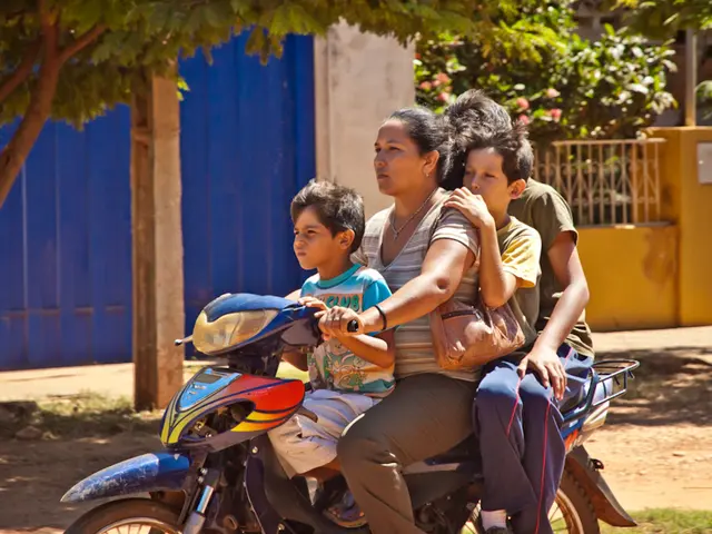 Here we can see a woman driving a motor cycle where a boy sitting in front and there are other two...