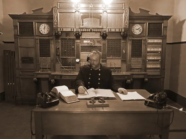 There is a person sitting writing something on a book with a pen behind him there is a telephone...