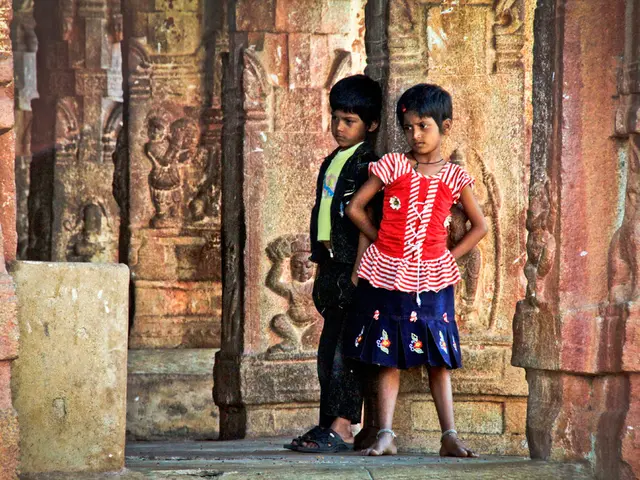 In this image two children are standing and one of the girl is wearing red color dress,and a boy is...