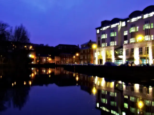 In this image in front there is water. In the background there are trees, buildings, street lights...