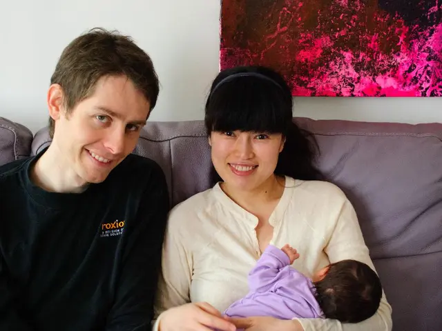 Woman in a white shirt who is holding a baby in her hand is sitting on the sofa and she is smiling....