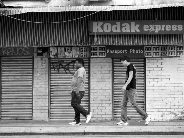 In the picture I can see two men are walking on the ground. In the background I can see shops,...