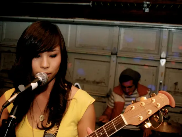 The picture is is of a live performance. In the foreground a woman is playing guitar and singing....