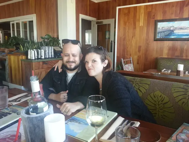 A man and a woman are sitting on the sofa. There are wine glasses,bottles on the dining table...