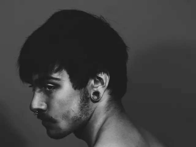 This is a black and white image, there is a man standing without shirt with earrings and nose ring.