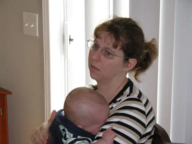 In this image there is a woman sitting on the chair. She is holding an infant in her hand. Behind...