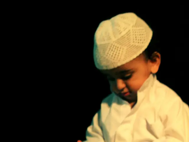 In this picture I can observe a kid sitting in front of a book. He is wearing a white color dress...