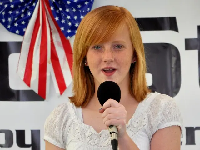 In this image we can see a lady standing and speaking into a microphone. There is a USA flag and a...