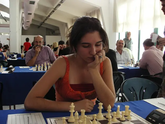 The women wearing a red dress is playing chess which is placed on a table with a blue mat on it and...