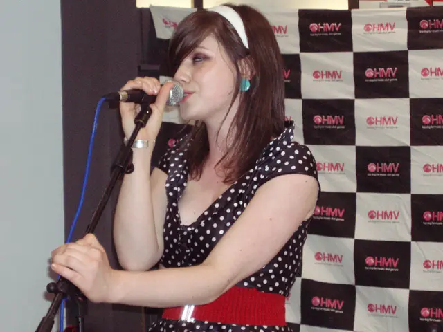 This image consists of a woman. She is wearing black color dress, there is a mic in front of her,...