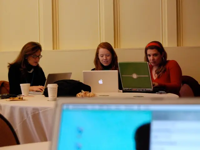 Three Women are sitting on the chairs around the round table and working in their laptops behind...