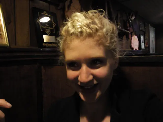 The woman in the front of the picture wearing black dress is smiling. Behind her, we see a wall on...