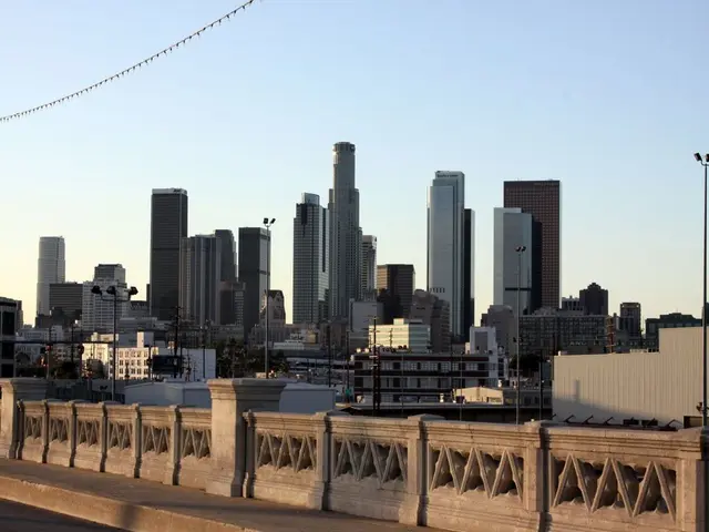 In the image there is a railing and behind the railing there are many tall buildings and poles.