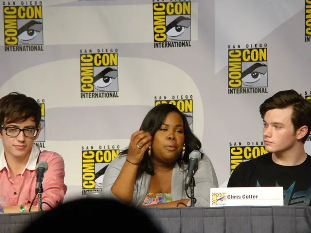 This picture shows a couple of men and women seated and we see microphones on the table and a name...