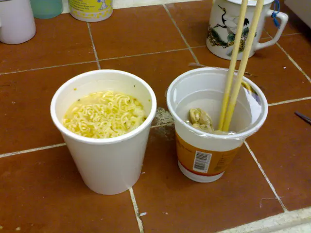 In the image there is a cup of noodles and beside it there is cup with chopsticks in it ,it on the...