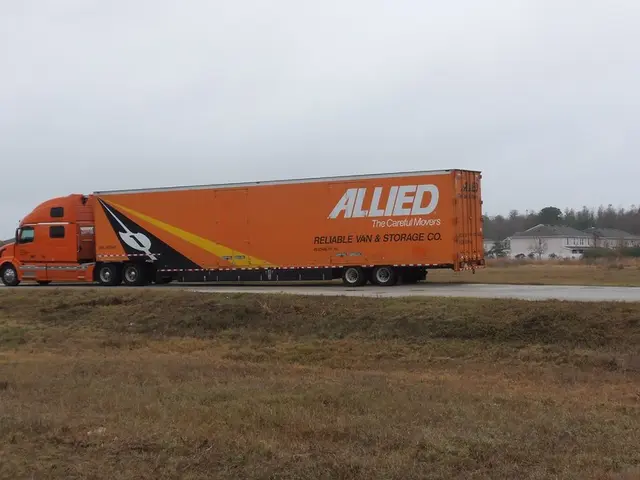 A big truck is moving on the road, it is in orange color, on the right side there are houses. At...