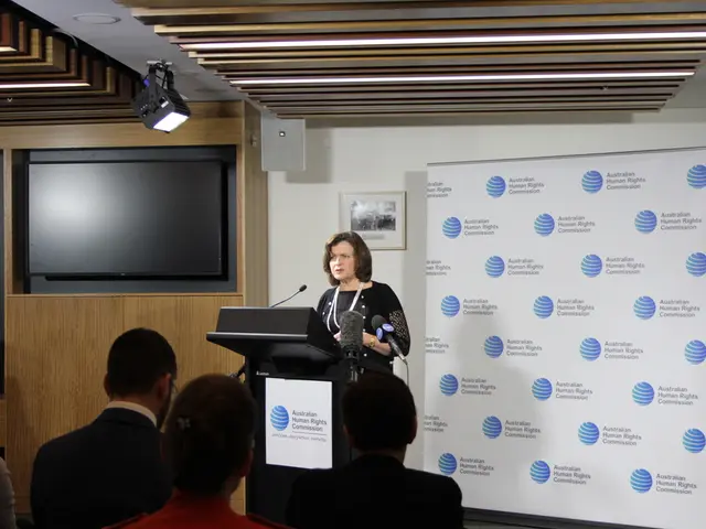 In this image there is a lady standing on the stage in front of table and speaking in a microphone,...