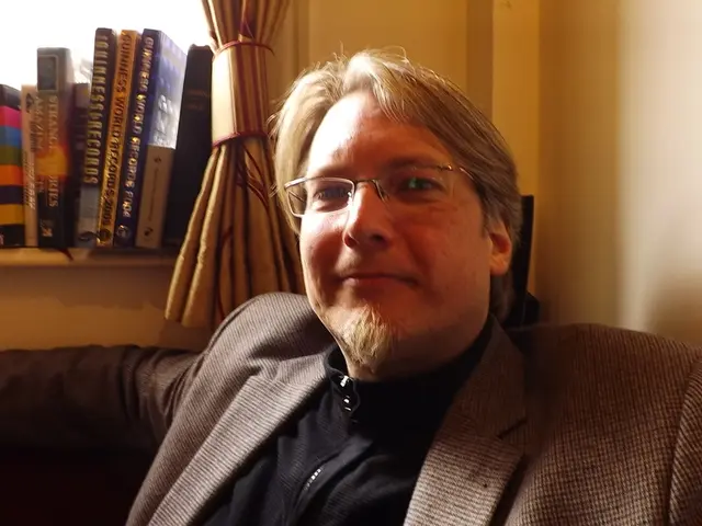 In the image we can see a man wearing clothes, spectacles and he is smiling. There are books kept...