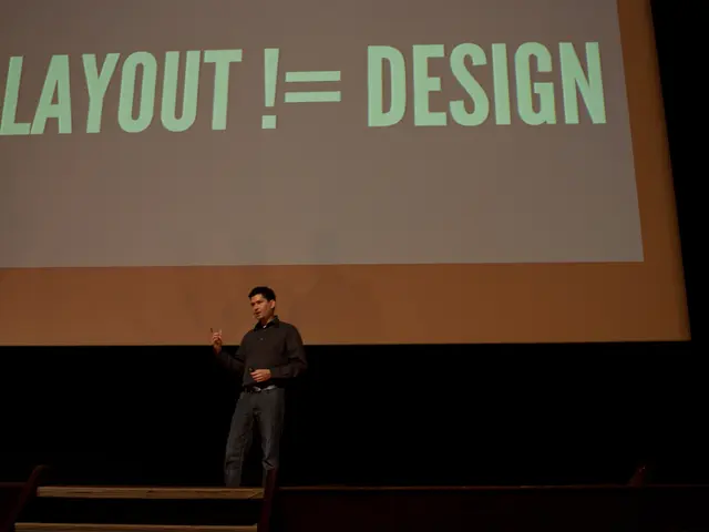 In this picture there is a person standing on the stage and talking. At the back there is a screen...