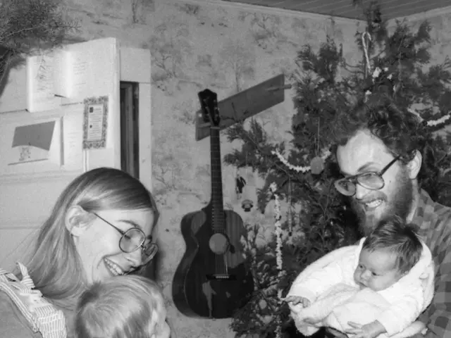 This image has a person holding a baby in his arms at the left side there is a woman wearing a...