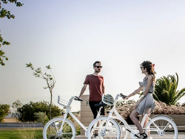 A man wearing goggles is holding a cycle and standing. A lady is sitting on a cycle. And the cycles...