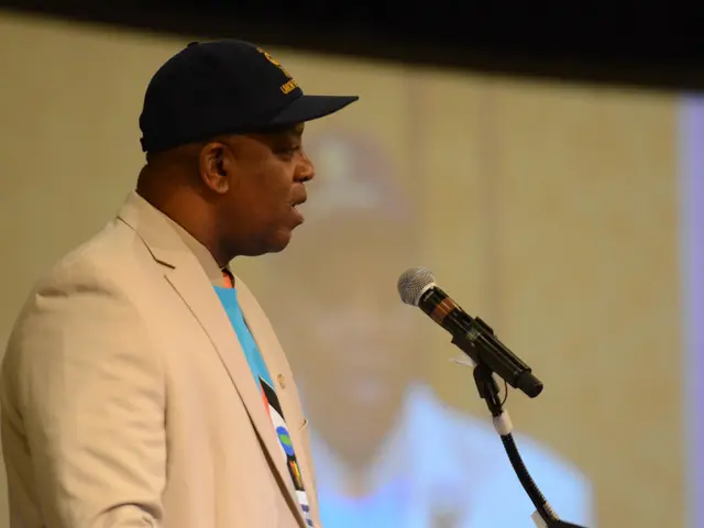 This picture is mainly highlighted with a man. He is wearing a black cap. He is standing and giving...